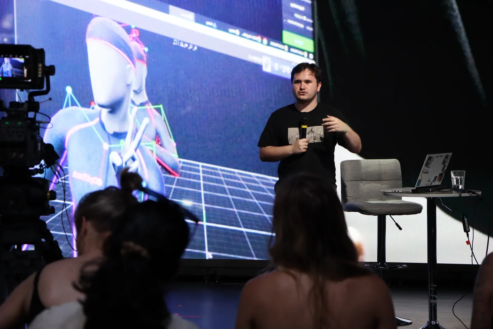 Joshua Prasser presenting in front of an audience with a 3D motion-capture avatar wireframe displayed on a virtual production LED wall.