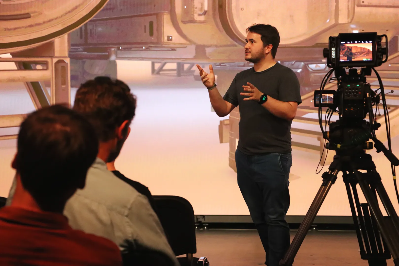 Joshua Prasser giving a live demonstration of virtual production technology to an audience in front of a large LED wall displaying a sci-fi environment, with a studio camera in the foreground.